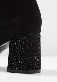 Bottine en daim noir avec un talon épais recouvert de petits strass noirs brillants, photographiée sur fond blanc.