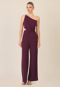 Jumpsuit viola con spallina unica, design frontale intrecciato, lati con apertura e gambe ampie, con un accento metallico sulla spalla.