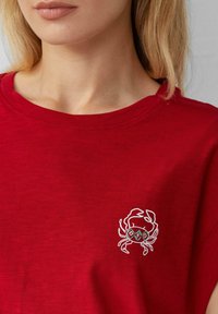 Vrouw in een rood shirt met een kleine gestileerde krab graphic in wit en teal op de linkerkant van de borst.
