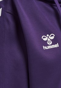 Felpa sportiva viola con texture liscia, logo Hummel bianco e accenti bianchi a contrasto lungo le cuciture laterali.