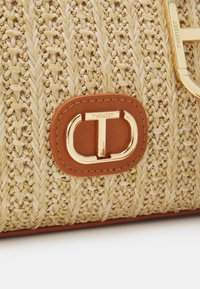 Kudottu olkalaukku oljenvärisessä sävyssä, jossa on vaaleanruskeat nahkaosat. Varustettu näyttävällä kultasävyisellä logovarusteella. Teksturoitu pinta, jossa on pystysuoria kuvioita.