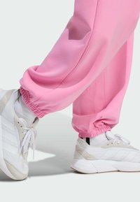 Ružové teplákové nohavice s elastickými manžetami v kombinácii s bielymi teniskami s plastickým šedým akcentom a prúžkovým vzorom po strane. Mäkká textúra látky.
