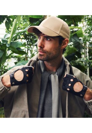 Man wearing beige cap, taupe jacket, gray scarf, and black fingerless gloves, standing among large green leafy plants.