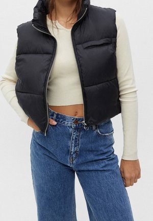 Frau trägt eine schwarze Steppweste über einem cremefarbenen langärmeligen Crop-Top und hochgeschnittene blaue Jeans, steht mit einer Hand in der Tasche.