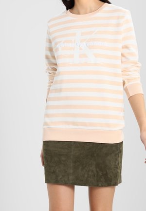 Kobieta w jasnopomarańczowej i białej, w paski bluzie Calvin Klein z dużym logo, zestawiona z oliwkową zamszową mini spódnicą.