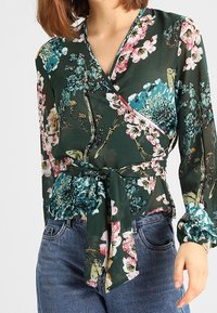 Blommig omslagsblus i mörkgrön, med rosa, blå och vita blommor. Långa ärmar med knytning i midjan; genomskinligt, lätt material.