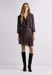 Il vestito presenta un tessuto scuro con righe verticali multicolori, maniche lunghe, collo con colletto e un orlo asimmetrico. Abbinato a stivali neri lucidi al ginocchio.