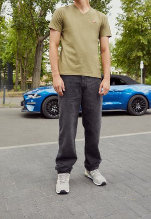 Persoon draagt een olijfgroen t-shirt, zwarte spijkerbroek en witte sneakers, staand op een stoep met op de achtergrond een blauwe cabriolet.