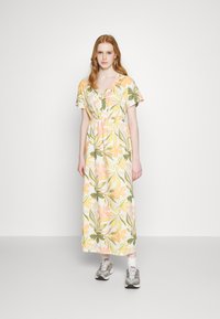 Fiatal nő virágos, rövid ujjú maxiruhában áll, tornacipőben, sima fehér háttér előtt.