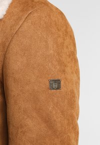 Brun mockaskinnjacka med axeln synlig, en liten fyrkantig metaltagg märkt "JEANSCODE" och en delvis vy av en vit fårskinnskrage.