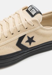 Vaaleanbeiget suedesneakerit mustalla kumipohjalla, jossa on musta tähti-logo ja nauhoitus. Siinä on myös musta logopläntti kielekkeessä.