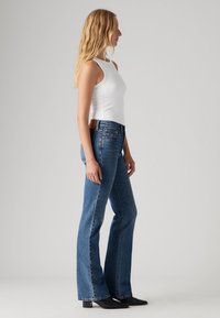 Kvinne med langt blondt hår iført en hvit ermeløs topp, blå high-waisted flared jeans og svarte høyhælte ankelstøvler, stående sidelengs.