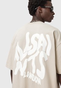 Young man wearing beige T-shirt with large white distorted text "ALSAINTS LONDON" on the back, sunglasses, and chain necklace.
