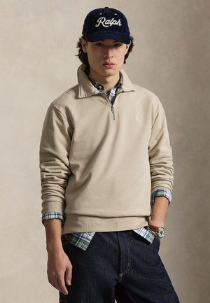 Ung mand iført en beige quarter-zip sweatshirt over en ternet skjorte, mørke jeans, en navyblå "Ralph" kasket og et armbåndsur med brun læderrem.