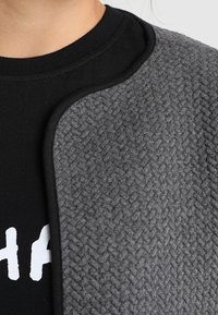 Tessuto grigio a motivo spigato con una texture liscia, bordato in nero. Indossato sopra una maglietta nera con lettering bianco.