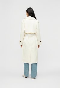 Femme portant un long trench-coat crème avec ceinture, un jean large bleu et des chaussures de couleur claire, debout de dos sur un fond uni.