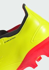 Scarpa sportiva gialla brillante con tomaia testurizzata, rivestimento interno nero, suola rossa e branding "HYBRIDFEEL". Presenta un design aerodinamico.