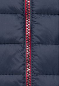 Tamsiai mėlynas pūkinis džemperis, turintis ryškų užtrauktuką su raudonais akcentais ir tekstūruota paklodė. Siuvinėta detalė su lygiu, lengvu audiniu.