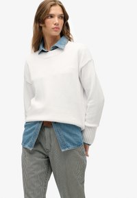 Joven mujer vistiendo un suéter blanco sobre una camisa de mezclilla azul y pantalones a rayas, mirando hacia un lado con un fondo neutro.