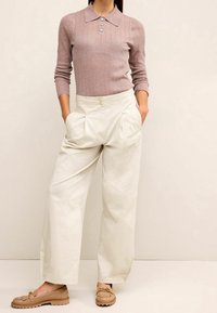 Vrouw in een mauve ribbelige polo trui met lange mouwen en knoopdetails, witte wijde broek en beige instappers met franjes en kettingaccenten.