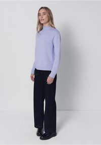 Hellvioletter Strickpullover mit hohem Kragen, kombiniert mit dunklen, weit geschnittenen Hosen und schwarzen Schuhen, vor einem schlichten Hintergrund.