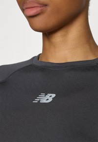Sivina športna majica z gladko teksturo, okroglo ovratnikom in odbojnim belim logotipom New Balance spredaj.