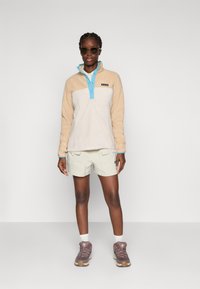 Tanskleurige fleecepullover met een blauwe kraag en knoopdetails, gecombineerd met licht khaki shorts en bruine wandelschoenen, die een ontspannen pasvorm laat zien.