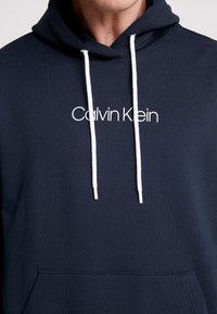 Námořnická modrá mikina s kapucí, s přední kapsou ve stylu klokaní, bílými šňůrkami a bílým logem "Calvin Klein" na hrudi v jednoduchém písmu.