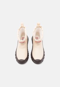 Fény bézs színű, fényes bevonatú bokacsizma, elasztikus felsőrésszel, fekete gumitalppal és piros címkével. Kerek orrú, barázdált talpú a jobb tapadás érdekében.