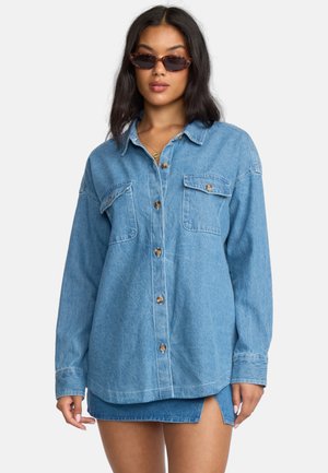 Ljusblå denimskjorta med långa ärmar, två bröstfickor, bruna knappar, avslappnad passform och en slät textur. Bärs över en matchande denimkjol.