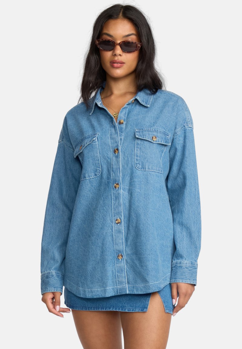 Light blue denim shirt with long sleeves, two chest pockets, brown buttons, relaxed fit, and a smooth texture. Worn over a matching denim skirt.
