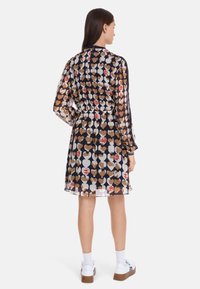 Das Kleid hat einen dunklen Hintergrund mit einem Herzmuster in Beigetönen, Weiß und Rot. Lange Ärmel sowie eine taillierte Taille; fließender Stoff.