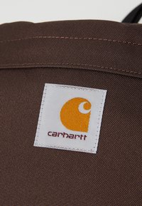 Braune Stofftasche mit einem quadratischen weißen Etikett, auf dem ein gelbes Logo und roter Text mit der Aufschrift "carhartt" zu sehen sind. Die Oberfläche wirkt strapazierfähig und strukturiert.