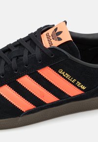 Musta mokkanahkainen tennari oransseilla ja mustilla raidoilla, jossa kultaiset sanat "GAZELLE TEAM". Kumipohja, jossa on kuvioitu pinta, klassinen muotoilu.