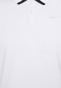 Bílá polo košile se strukturovanou látkou, černým límečkem, tříknoflíkovým průkrčníkem a stříbrným logem Nike na horní levé straně.