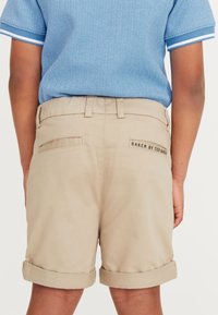 Khaki shorts met opgerolde manchetten, voorzien van twee achterzakken en een label met de tekst "BAKER BY TED BAKER." Glad stoftextuur.