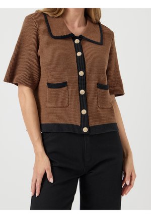 Cardigan en maille marron à manches courtes avec bordure noire, boutons dorés, deux poches avant, porté avec un pantalon noir.