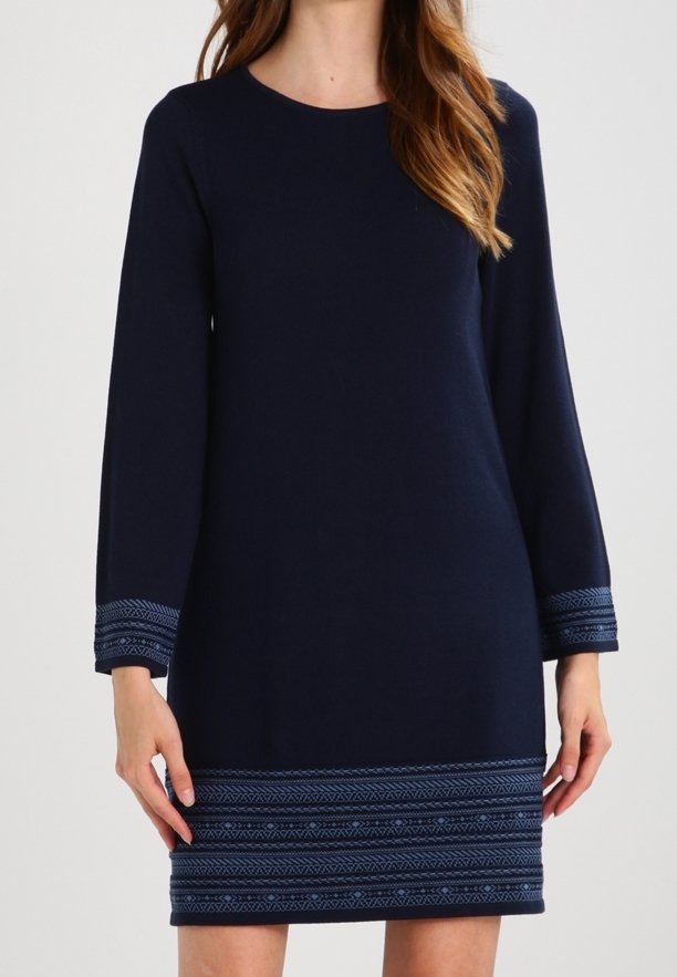 Femme portant une robe marine longueur genou avec des manches longues et des bandes bleues à motifs aux poignets et à l'ourlet, debout devant un fond uni.