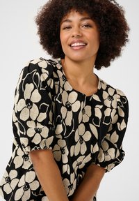 Blouse noire avec un motif floral crème, des manches bouffantes, un col rond et une coupe décontractée. Le tissu présente des détails texturés.