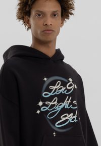 Zwarte hoodie met een voorzak heeft een grafisch ontwerp met blauwe en witte tekst, omringd door sterren op een zachte stoftextuur.