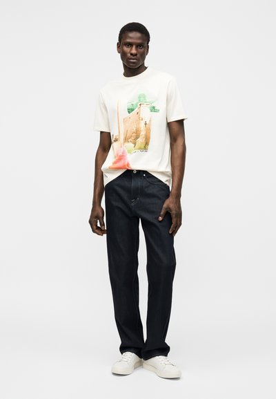 Mees, kes kannab valget graafilist T-särki Egiptuse trükiga, tumedaid teksapükse ja valgeid tossusid, seisab ühevärvilise heleda tausta ees.