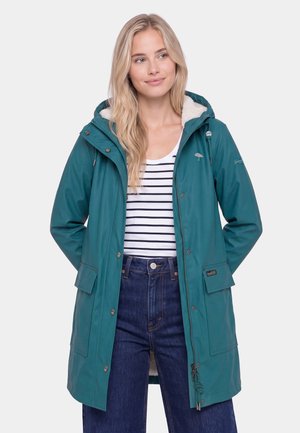 Frau trägt eine türkisfarbene Kapuzenregenjacke über einem weiß-navy gestreiften Hemd und dunkelblauen High-Waist-Jeans, Hände hinter dem Rücken, schaut leicht nach rechts.