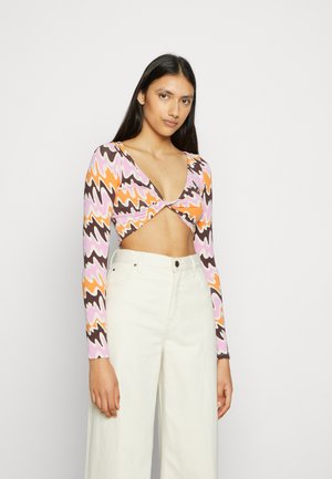 Top cropped de manga longa em padrão de ondas rosa, laranja e marrom, com um decote em V profundo e detalhe torcido na frente. Emparelhado com calças de perna larga de cor clara.
