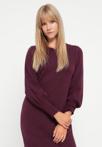 Burgundy kötött ruha hosszú, enyhén puffos ujjal és bordás mandzsettával. Az anyaga lágy és sima megjelenésű, formázott sziluettel rendelkezik.
