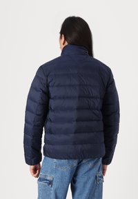Geacă puffer de culoare bleu marin, cu guler înalt, secțiuni matlasate și manșete elastice. Proiectată pentru căldură, având un exterior ușor și neted.