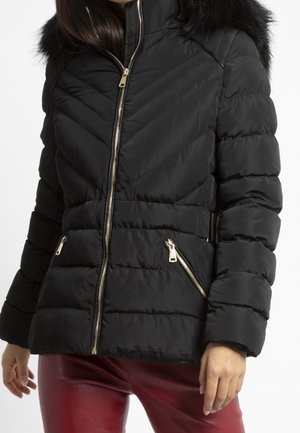 Femme portant une veste d'hiver matelassée noire avec une capuche bordée de fourrure et des détails de fermeture éclair dorés, assortie à un pantalon en cuir rouge.