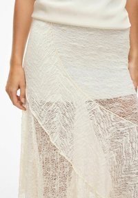 Jupe en dentelle de couleur blanc cassé présentant des textures superposées, un motif en zigzag et une finition semi-transparente. Comprend une taille cintrée et une silhouette fluide.