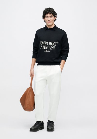 Czarna bluza z białym logo "EMPORIO ARMANI", kremowe spodnie, czarne trampki, trzyma torbę typu tote z brązowej skóry, gładka tekstura.