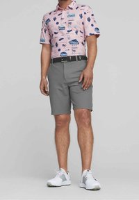 Polo de golf rose avec un cactus et un imprimé graphique, associé à un short gris. Des chaussures de sport blanches complètent la tenue.