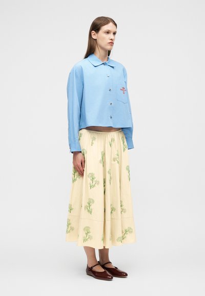 Γυναίκα που στέκεται, φοράει ανοιχτό μπλε πουκάμισο με κουμπιά, κρεμ φούστα με πράσινα λουλουδένια σχέδια και σκούρα καφέ παπούτσια Mary Jane σε απλό φόντο.